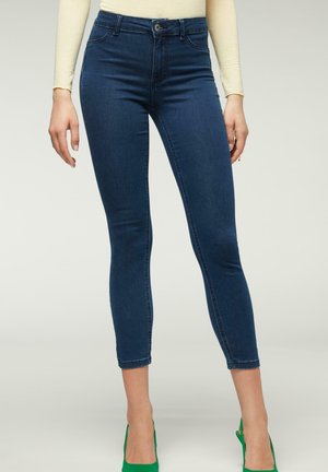 Vrouw draagt donkerblauwe skinny jeans, beige top met lange mouwen en groene hoge hakken, staand tegen een effen lichte achtergrond.