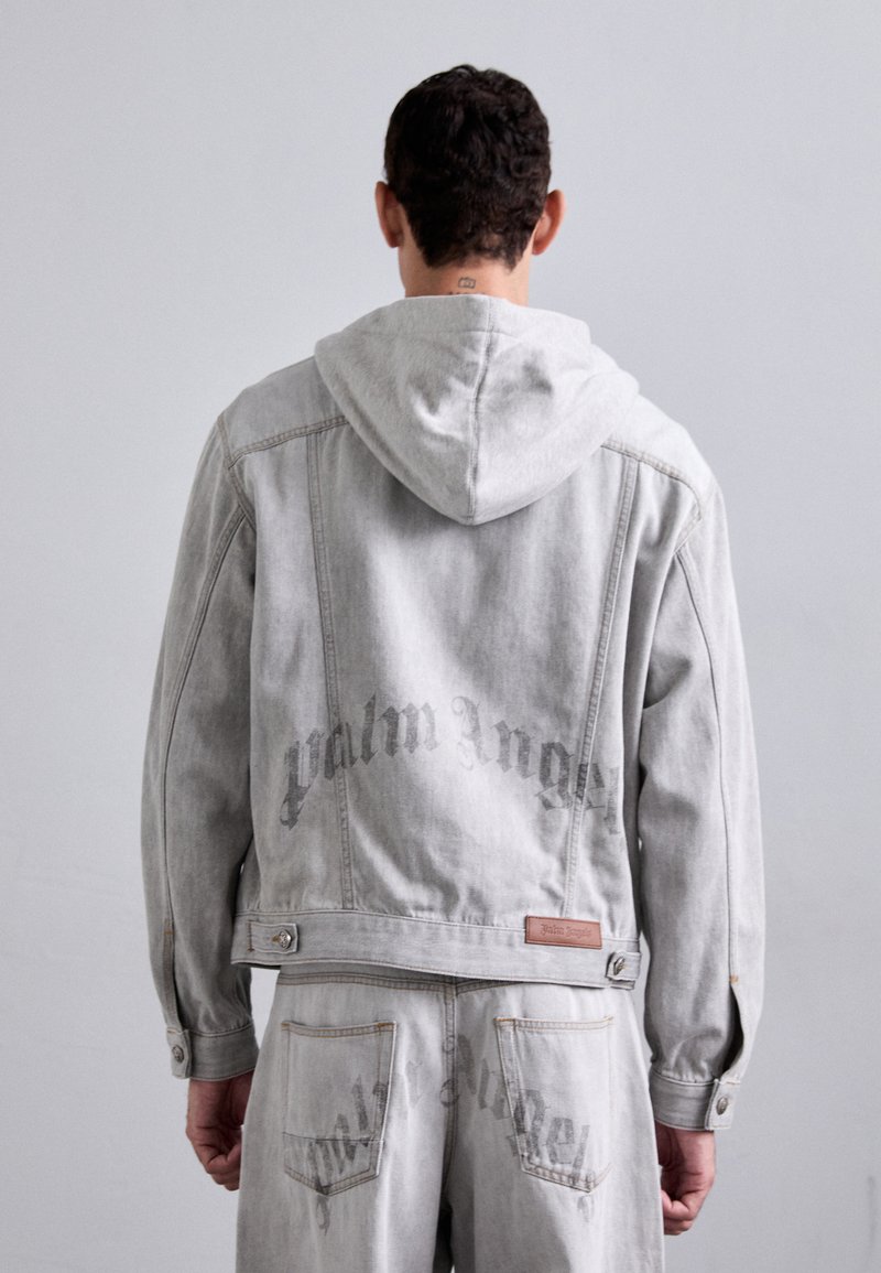 Grå denimjakke med hætte, synlige syninger og "Palm Angels" tekst på ryggen. Inkluderer en læderetiket og knapdetaljer.