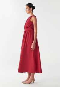 Punainen sleeveless-maxi-mekko, jossa on kootut vyötärö, sileä kangas ja virtaava hame. Yhdistetty beigen koristellun kengän kanssa.