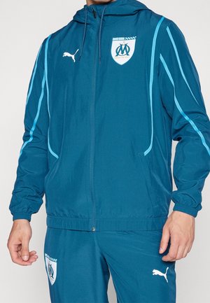 Teal windbreaker with hood, white Puma logo, and OM crest. Features blue side stripes, elastic cuffs, and zippered front pockets.