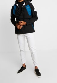 Czarny i niebieski wiatrówka w kolorze blokowym z kapturem, kieszenią z przodu i elastycznymi mankietami, zestawiona z białymi zwężanymi spodniami i czarnymi sneakersami.