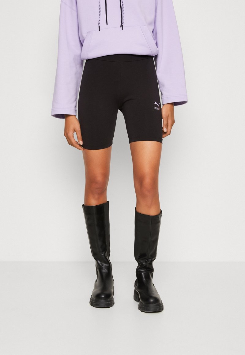 Personne portant un cuissard de cyclisme noir Puma, un sweat à capuche lavande et des bottes noires montant jusqu'aux genoux, debout devant un fond blanc uni.
