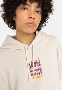 Crème hoodie met een voorkantgrafiek in blauwe, rode en gele tekst. Voorzien van een trekkoordcapuchon en geribde manchetten, gemaakt van zachte stof.
