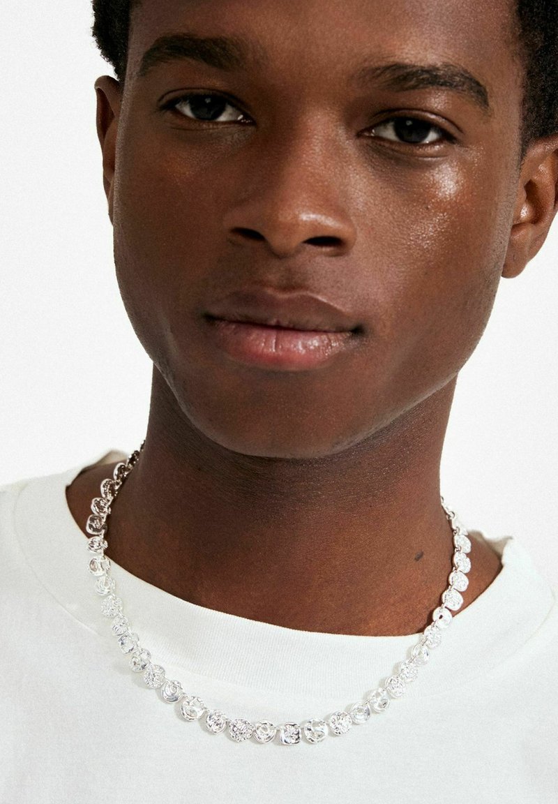 Collar de plata con cuentas redondas y texturizadas en varios patrones, llevado sobre una camiseta blanca, con piel lisa visible debajo.