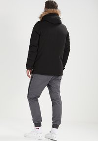 Svart parkas med pelsforet hette, kombinert med grå joggebukser. Begge har funksjonelle lommer, teksturert stoff og elastiske mansjetter for god passform.