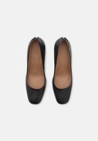 Escarpins noirs en cuir avec bout rond, présentant une texture lisse et un subtil marquage à l'intérieur. Détails dorés remarquables à l'arrière.
