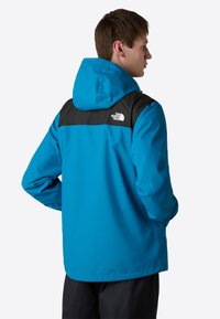 Modrá nepromokavá bunda s černými ramenními akcenty, kapucí a detailem loga. Má uvolněný střih a hladkou, texturovanou látku.