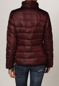 Veste matelassée bordeaux avec un col haut, un design matelassé et une finition brillante. Portée sur un jean bleu, mettant en avant les détails de couture et de texture.