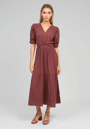 Femme portant une robe midi mauve portefeuille à manches bouffantes avec une jupe à volants et des sandales à talons blocs marron, debout sur un fond gris clair.