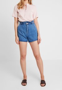 Mujer con una blusa corta de manga corta en rosa claro metida dentro de unos pantalones cortos de mezclilla azul de cintura alta con una cintura con volantes y sandalias de tiras trenzadas marrones.