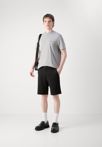 Un t-shirt gris clair à manches courtes avec impression, un short noir ajusté, des chaussettes blanches et des mocassins noirs sont portés par un mannequin.