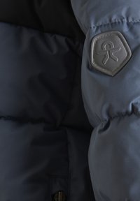 Blizu prikaz tamnoplave puffer jakne s teksturiranim, punjenim materijalom i heksagonalnom gumenom oznakom na rukavu.