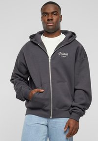 Gray zip-up hoodie with a kangaroo pocket, drawstring hood, and white embroidered "Prohibited" logo on the chest. Cotton blend material.