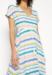 Femme portant une robe à manches courtes et col en V avec des rayures horizontales multicolores et un cordon de serrage blanc à pompons à la taille.