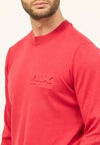 Maglione rosso a maniche lunghe realizzato in materiale morbido, caratterizzato da un colletto rotondo a coste e un logo in rilievo sul lato sinistro del petto.