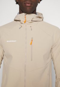 Veste beige à capuche avec des zips à l'avant et sur la poitrine, dotée de tirettes orange vif, portée par une personne recadrée du nez à la poitrine.
