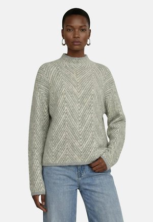 Kvinde iført en lysegrå strikket sweater med chevronmønster og lyseblå jeans, stående med den ene hånd i lommen mod en hvid baggrund.