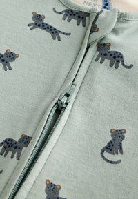 Vêtement zippé vert clair avec une texture douce, présentant un motif ludique de léopards bleus et gris avec des accents orange sur les têtes.