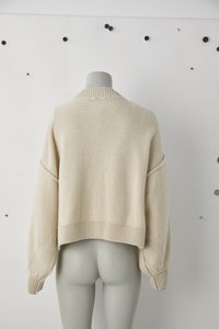 Cream-colored, cropped knit sweater with oversized sleeves, ribbed collar and hem. Subtle texture, relaxed fit, tag visible at the neck.