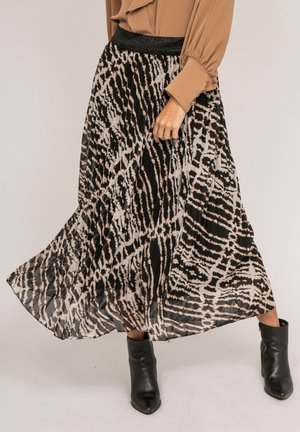 Femme portant une jupe plissée à imprimé animal noir et blanc, un chemisier beige et des bottines noires, debout devant un fond uni.