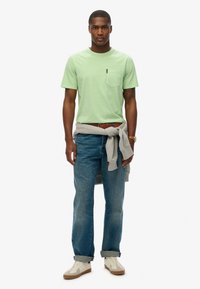 Lichtgroene katoenen T-shirt met een pocket, gecombineerd met blauwe denim jeans en een grijze trui om de taille. Off-witte sneakers maken de look compleet.