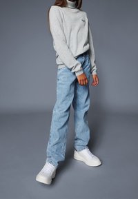 Suéter gris de cuello alto, pantalones vaqueros anchos de color azul claro y zapatillas deportivas blancas. El suéter tiene un pequeño logo; los pantalones son ligeramente descoloridos y de corte holgado.