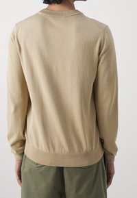 Beige neulepusero, jossa on pyöreä kaula-aukko, resorit hihansuissa ja helmassa, sekä sileä pinta ja vartalonmyötäinen muoto.