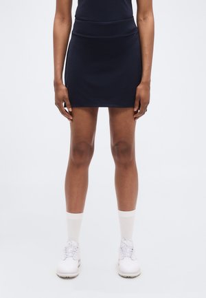 Σκούρα μπλε αθλητική φούστα-παντελόνι με λείο ελαστικό ζωνάρι στη μέση και εφαρμοστή σχεδίαση, συνδυασμένη με λευκά παπούτσια και κάλτσες μήκους μέχρι τον αστράγαλο.