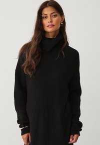 Sort strikket sweater med høj, foldet turtle neck og lange ærmer. Har et struktureret mønster og en tætsiddende silhuet.