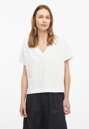Femme aux cheveux châtain clair portant un haut blanc ample à col en V et manches courtes ainsi qu’un pantalon noir taille haute à jambes larges, debout sur fond blanc.