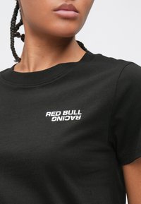 Κοντινό πλάνο ενός ατόμου που φοράει μαύρο T-shirt με λευκό κείμενο "RED BULL RACING" τυπωμένο κοντά στον αριστερό ώμο.