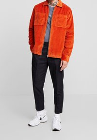 Giacca in corduroy arancione con due tasche sul petto, abbinata a una camicia grigia e pantaloni scuri. Indossata con scarpe da ginnastica bianche.