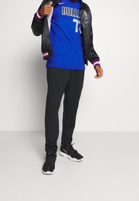 Uomo in giacca satin blu con jersey da basket di Dallas, pantaloni neri e sneakers nere con suola bianca.