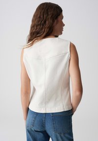 Weißes ärmelloses Oberteil aus Denim-Material, mit V-Ausschnitt, strukturierten Nähten und einem leicht verkürzten Design. Kombiniert mit blauen Jeans.