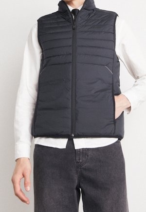 Homme portant un gilet matelassé noir zippé sur une chemise blanche avec une cravate noire et un jean foncé, main dans la poche du gilet.