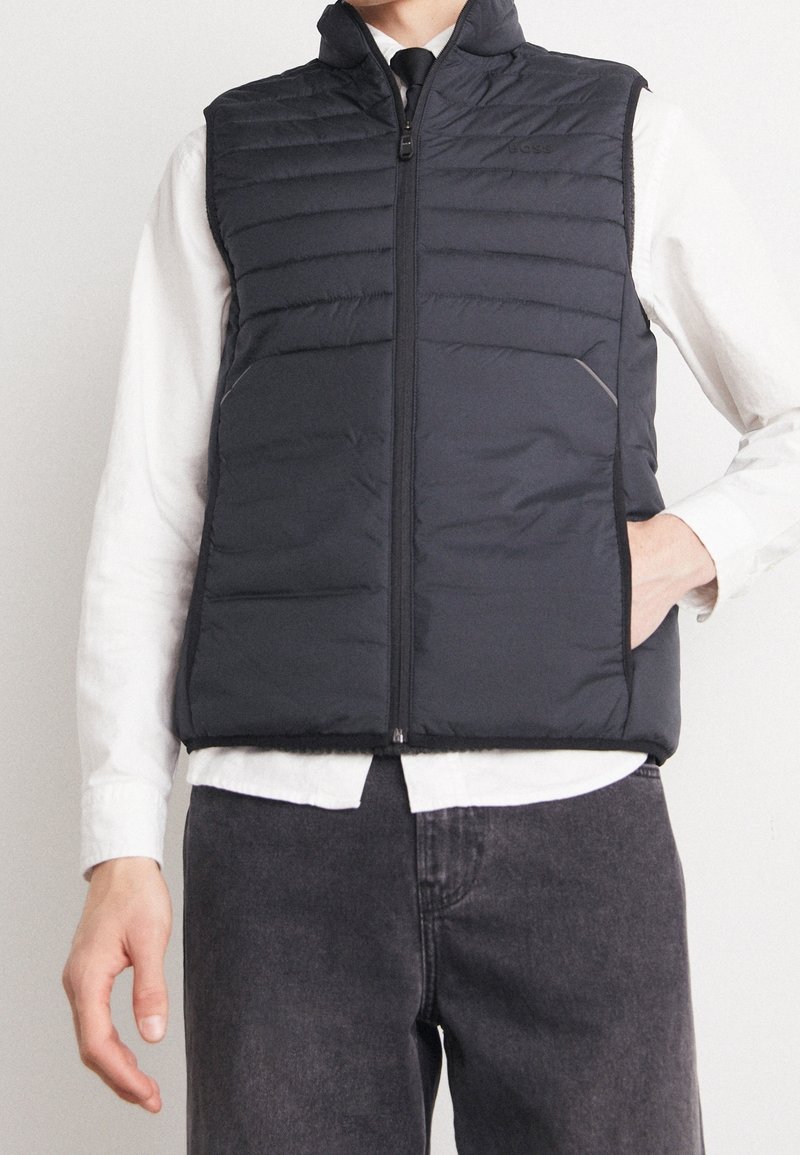 Homme portant un gilet matelassé noir zippé sur une chemise blanche avec une cravate noire et un jean foncé, main dans la poche du gilet.