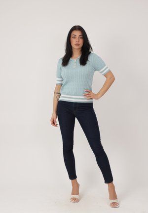 Donna in piedi con la mano sul fianco, indossa un maglione a maniche corte azzurro chiaro con bottoni, jeans skinny scuri e tacchi bianchi aperti sulle dita, su sfondo semplice.