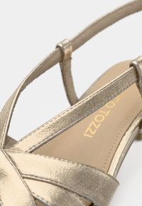 Kratak prikaz metalik zlatne sandale sa remenčićima i tankim remenom oko gležnja na neutralnoj pozadini.