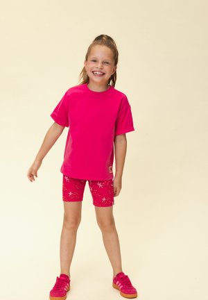 Lächendes Mädchen steht, trägt ein leuchtend pinkes T-Shirt, rote Shorts mit Erdbeermuster und pinke Turnschuhe vor einem einfarbigen beigen Hintergrund.