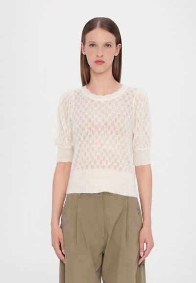 Pull crème tricoté en motif ajouré avec des manches bouffantes courtes et un décolleté à volants, associé à un pantalon taille haute vert olive.