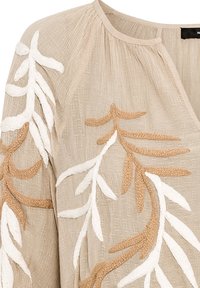 Blouse beige texturée avec un col rond décoré de motifs brodés en relief représentant des branches feuillues en blanc et beige.