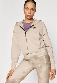 Μπεζ cropped φούτερ με μπροστινό φερμουάρ, ρυθμιζόμενη κουκούλα και λογότυπο. Φορεμένο με ανοιχτό καφέ κολάν με σχέδια και πλαϊνές τσέπες.