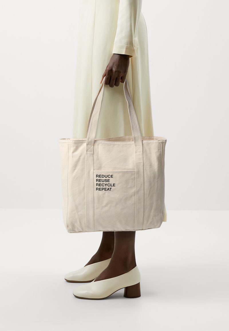 Canvas tote bag in light beige with a rectangular shape. Features "REDUCE REUSE RECYCLE REPEAT" in black text on the front.