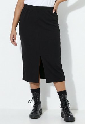 Femme portant une jupe noire mi-longue avec une fente à l'avant, des chaussettes noires aux chevilles et des bottines noires à lacets, debout contre un mur blanc.