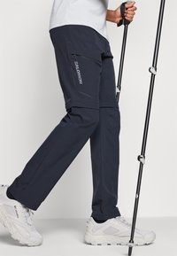 Pantalons de plein air bleu marine avec une poche zippée, dotés d'une coupe droite. Associés à des chaussures de randonnée grises et un bâton de trekking.