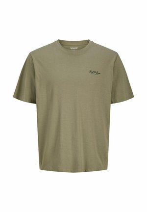 Tienerjongen staand, draagt een olijfgroene T-shirt, zwarte broek en beige sneakers, kijkt iets opzij tegen een witte achtergrond.
