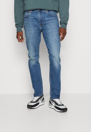 Mand iført blå tætsiddende jeans, sorte og hvide sneakers, og en grøn sweatshirt, stående mod en ensfarvet hvid baggrund.