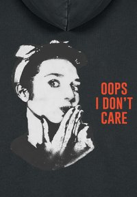 Fejkendőt viselő nő, aki kezeit a szája közelében tartja, oldalra néz, mellette vastag piros felirat olvasható: "HÚ, ÉN TÉNYLEG NEM ÉRDEKEL" sötét szövetes háttéren.