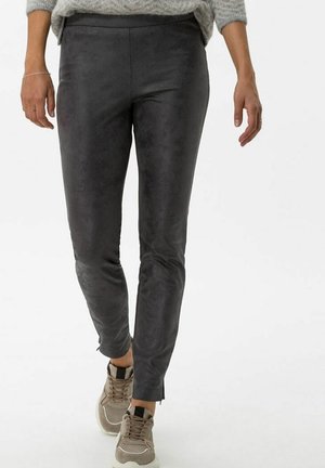 Personne portant un pantalon gris foncé ajusté et des baskets beiges, debout avec une jambe légèrement en avant sur un fond uni.