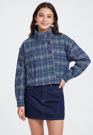 Junge Frau trägt eine blaue karierte, hochgeschlossene Jacke mit Knöpfen und einen dunklen Jeansrock, lächelt mit einer Hand in der Hüfte.